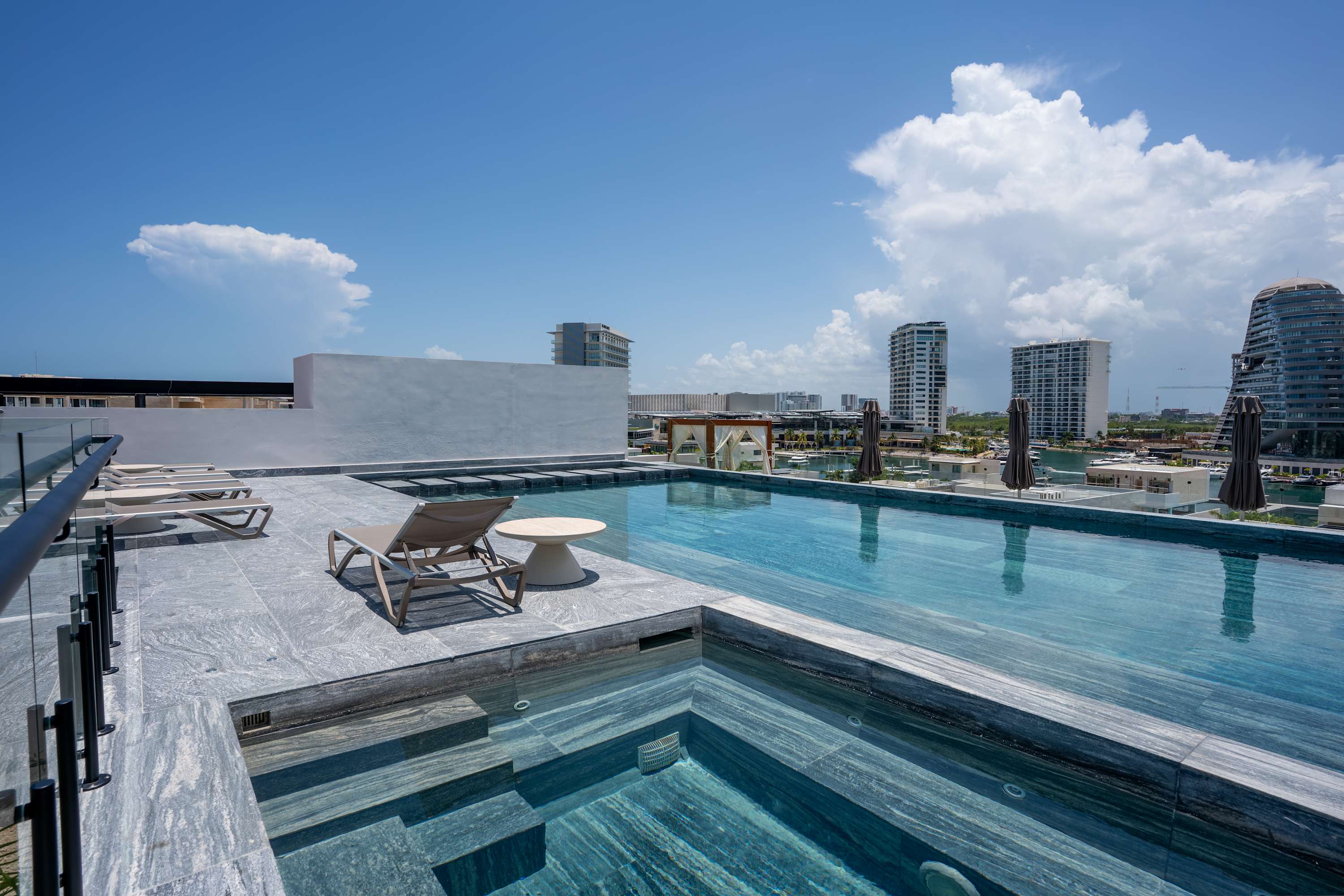 A rooftop pool with lounge chairs overlooks a city skyline and ocean under a blue, cloudy sky.