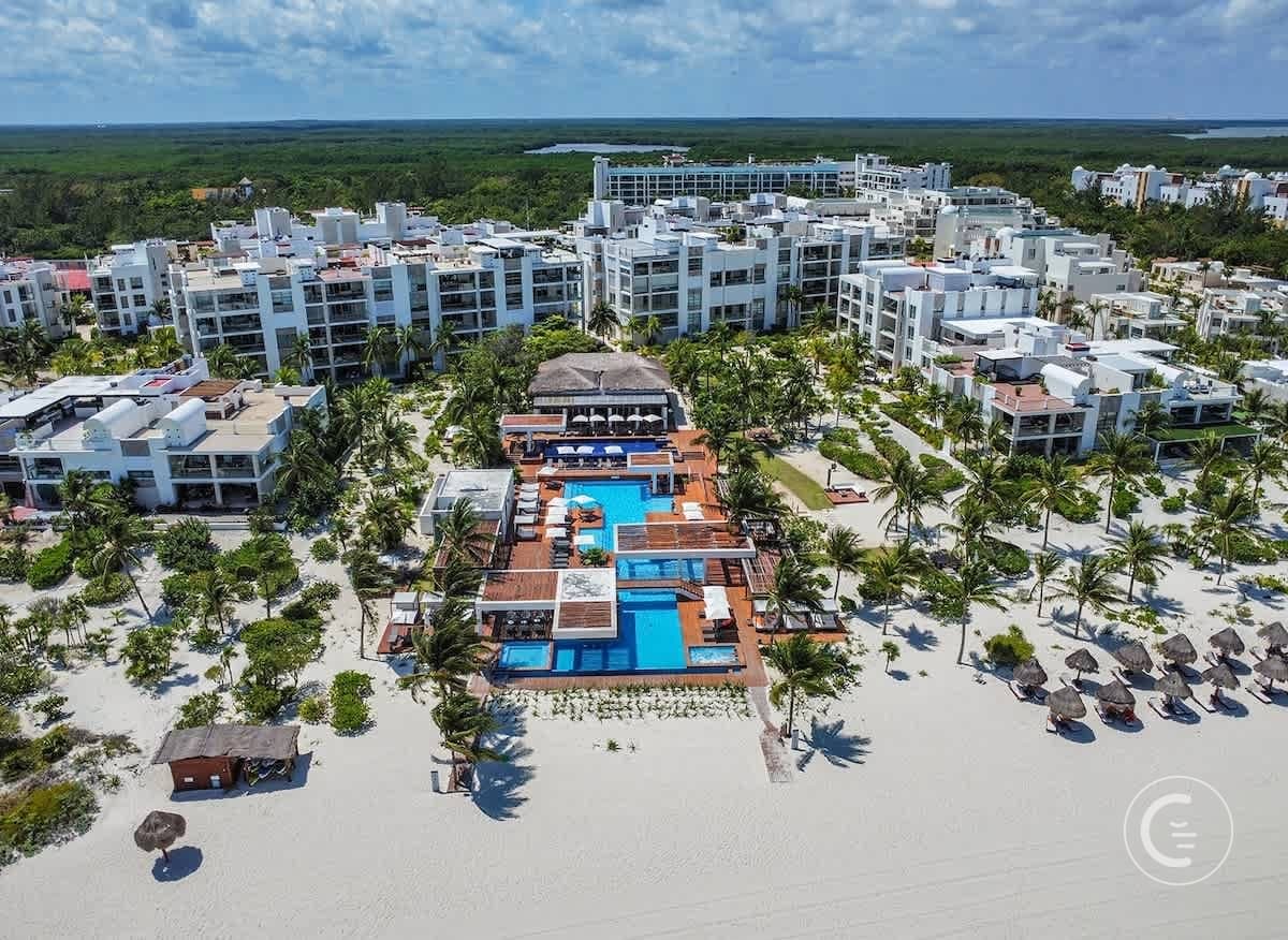 Aerial view of a resort with white buildings, pools, palm trees, and a sandy beach.