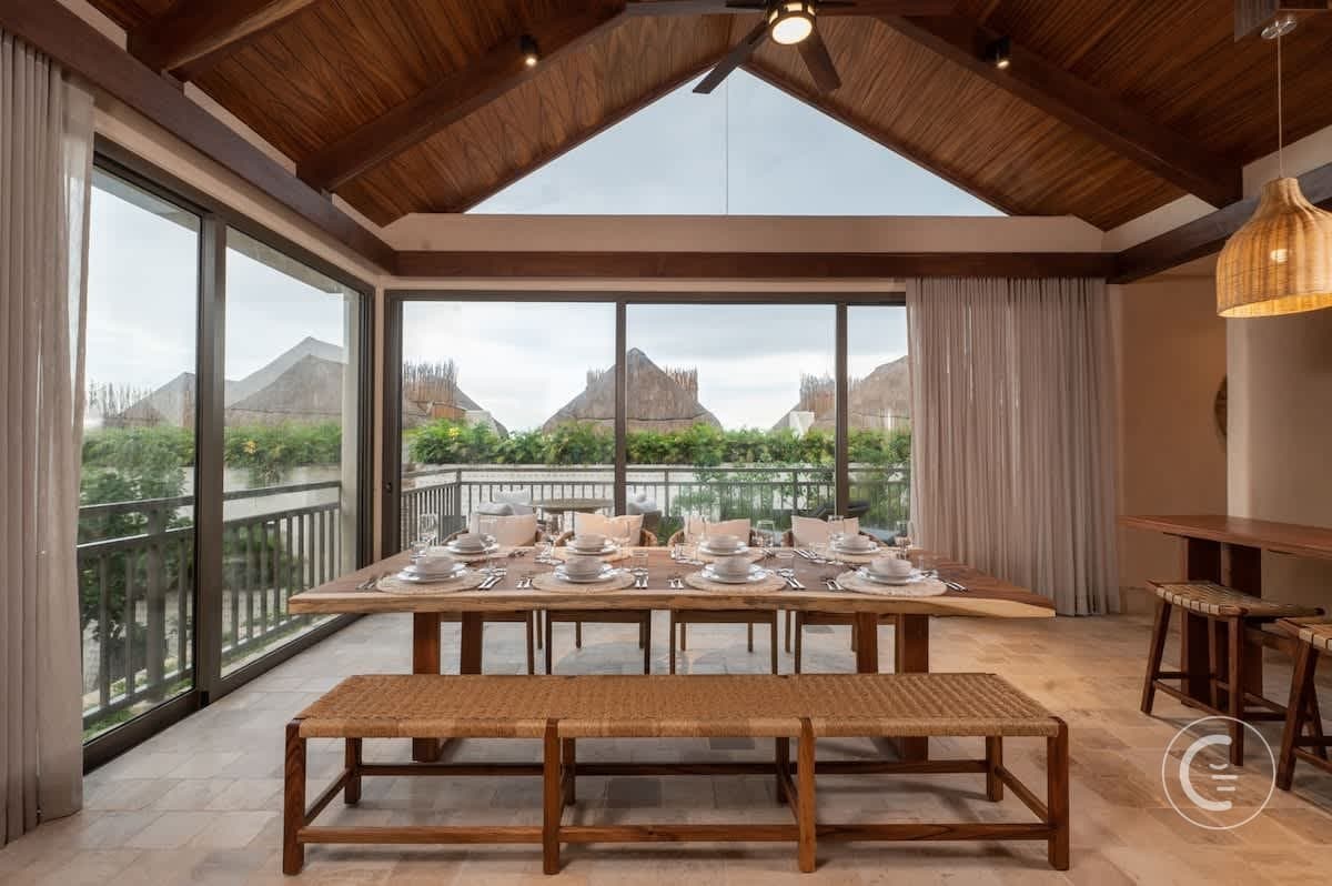 A dining room with a rustic wooden table set for a meal, overlooking a balcony with thatched-roof bu