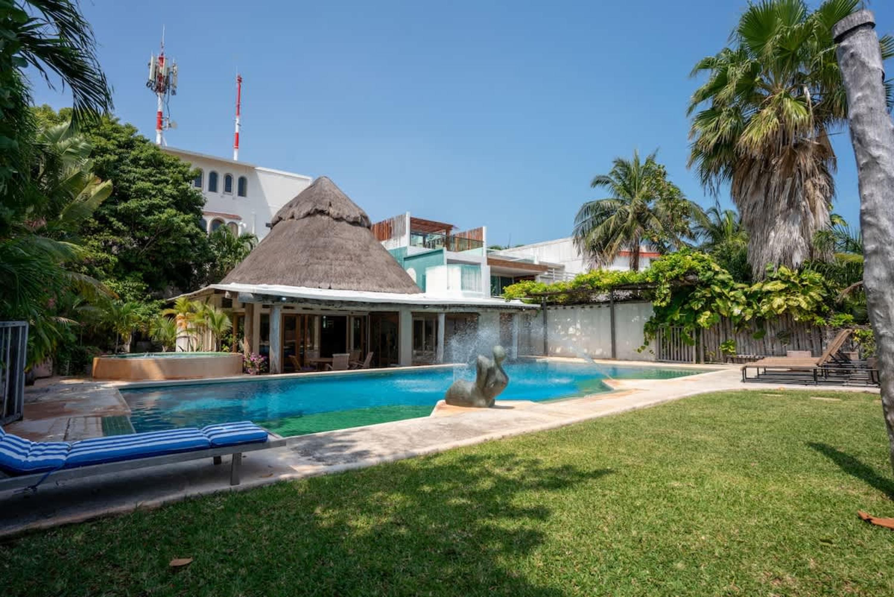 A resort-style swimming pool with a thatched-roof cabana, lounge chairs, and palm trees.