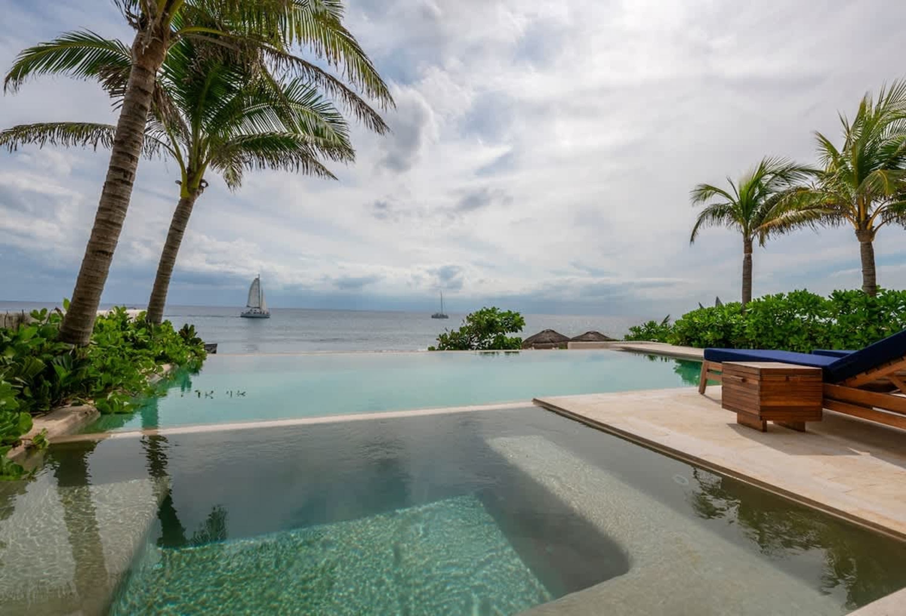 Infinity pool overlooking a calm ocean with sailboats and palm trees.